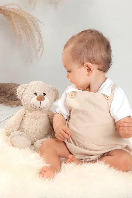 Baby in beige tuinbroekje zit op een wit vachtje naast een grote teddybeer – babyfotoshoot met knuffel in Hasselt