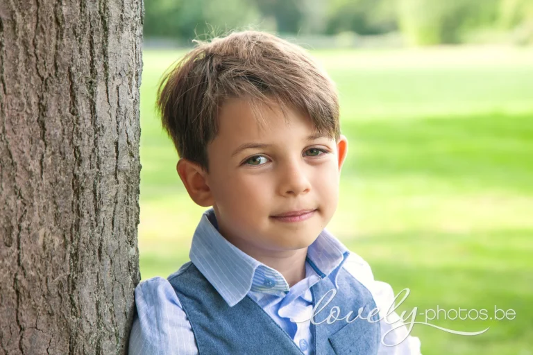 Jongen in lichtblauw hemd en blauw gilet leunt glimlachend tegen een boomstam in een groen park – communie fotoshoot op locatie in Limburg