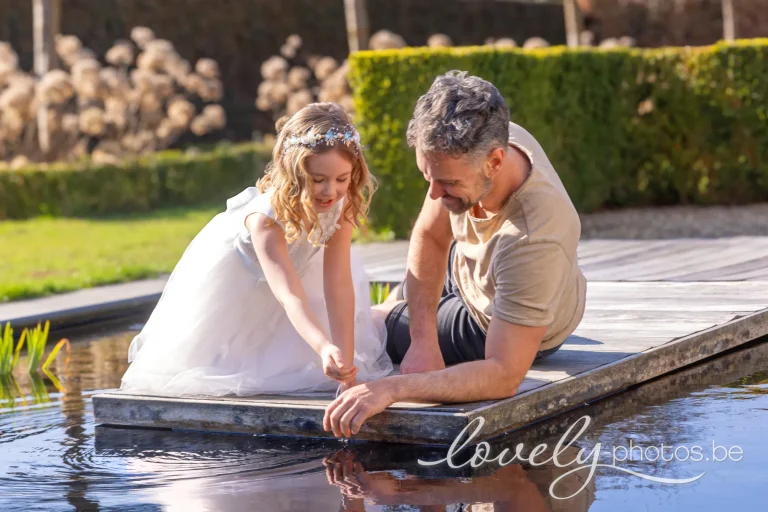 Communiemeisje in witte tule jurk met bloemenkroontje speelt lachend met het water van de zwemvijver, samen met haar papa op het houten vlonder – communie fotoshoot in de tuin van Lovely-Photos in Hasselt