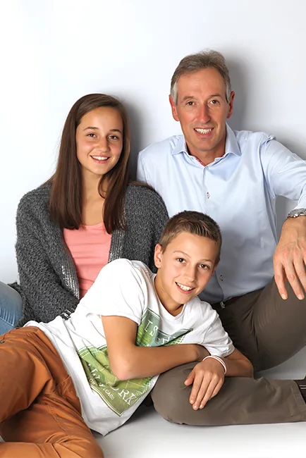 Vader zit samen met zijn tienerdochter en zoon lachend op de grond in de lichte fotostudio – familiefotoshoot in Hasselt