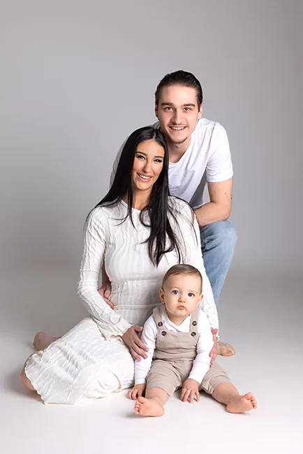 Jonge ouders zitten samen met hun babyzoon in beige salopette op een lichtgrijze achtergrond – Baby & Family fotoshoot bij Lovely-Photos in Hasselt