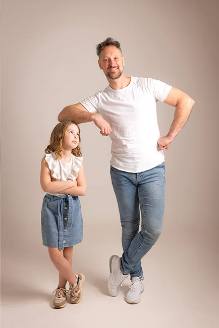 Vader in wit T-shirt en jeans leunt speels met zijn arm op het hoofd van zijn dochter in jeansrokje – family fotoshoot met bijpassende outfits in Hasselt