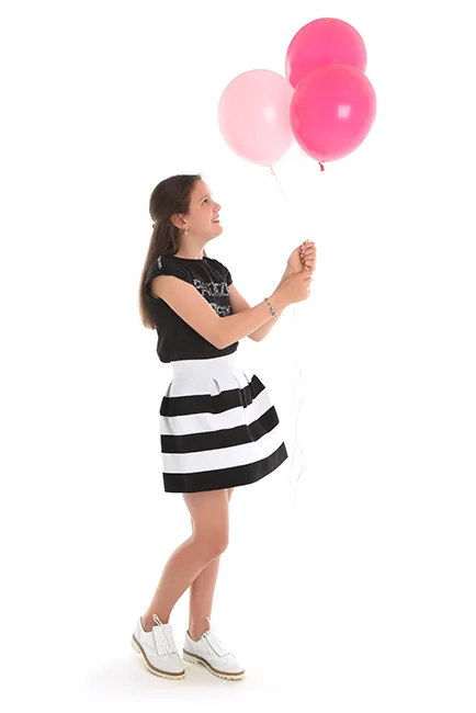 Tienermeisje in zwart T-shirt en zwart-wit gestreepte rok kijkt lachend naar drie roze ballonnen tegen een witte achtergrond – lentefeest- en communiefotoshoot in de studio in Hasselt
